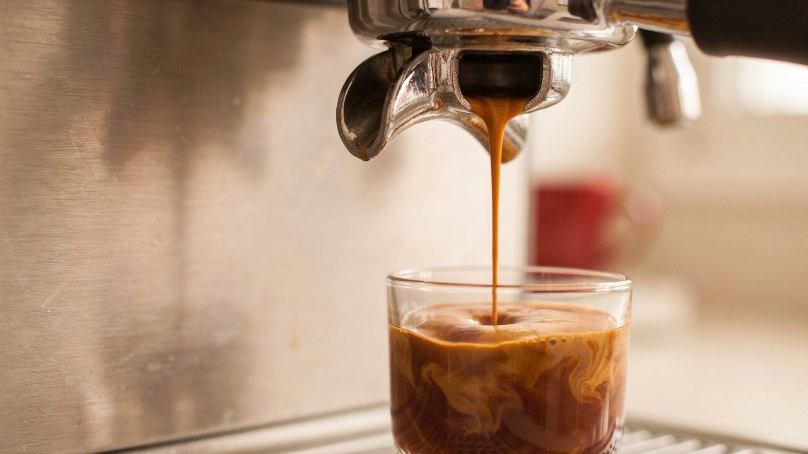 A bottomless (naked) portafilter on a brushed-steel home espresso machine, a single thick stream of espresso flowing from the center of the basket into a small glass, rich tiger-striped crema
