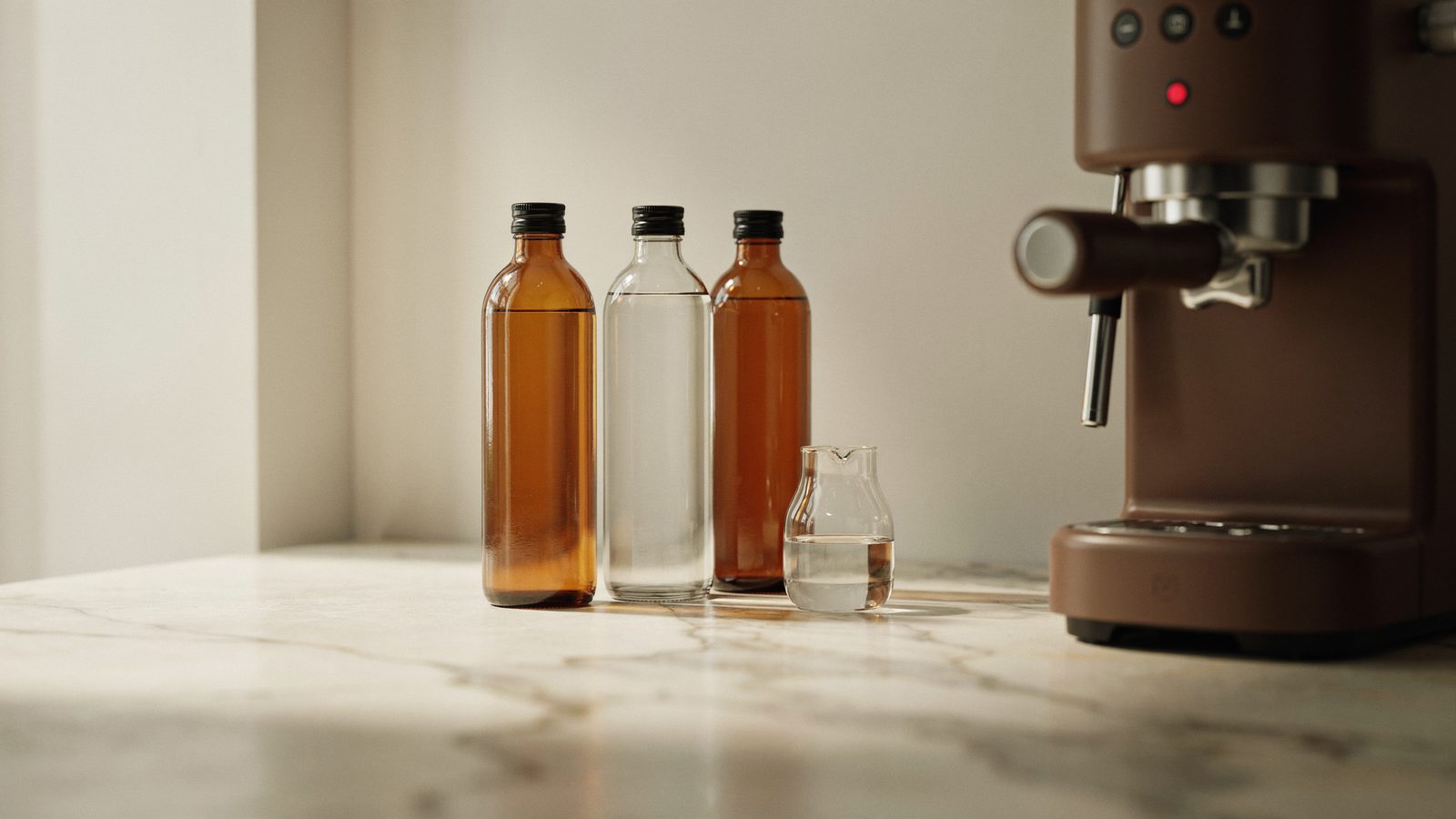 A still life of three plain unlabeled bottles of liquid descaler in amber and clear glass beside a small glass carafe of water on a marble counter next to the side of a home espresso machine