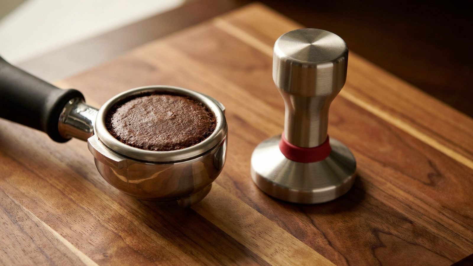 A stainless steel flat espresso tamper resting beside a portafilter filled with an evenly tamped, perfectly level coffee puck on a walnut tamping mat, close-up from a low angle