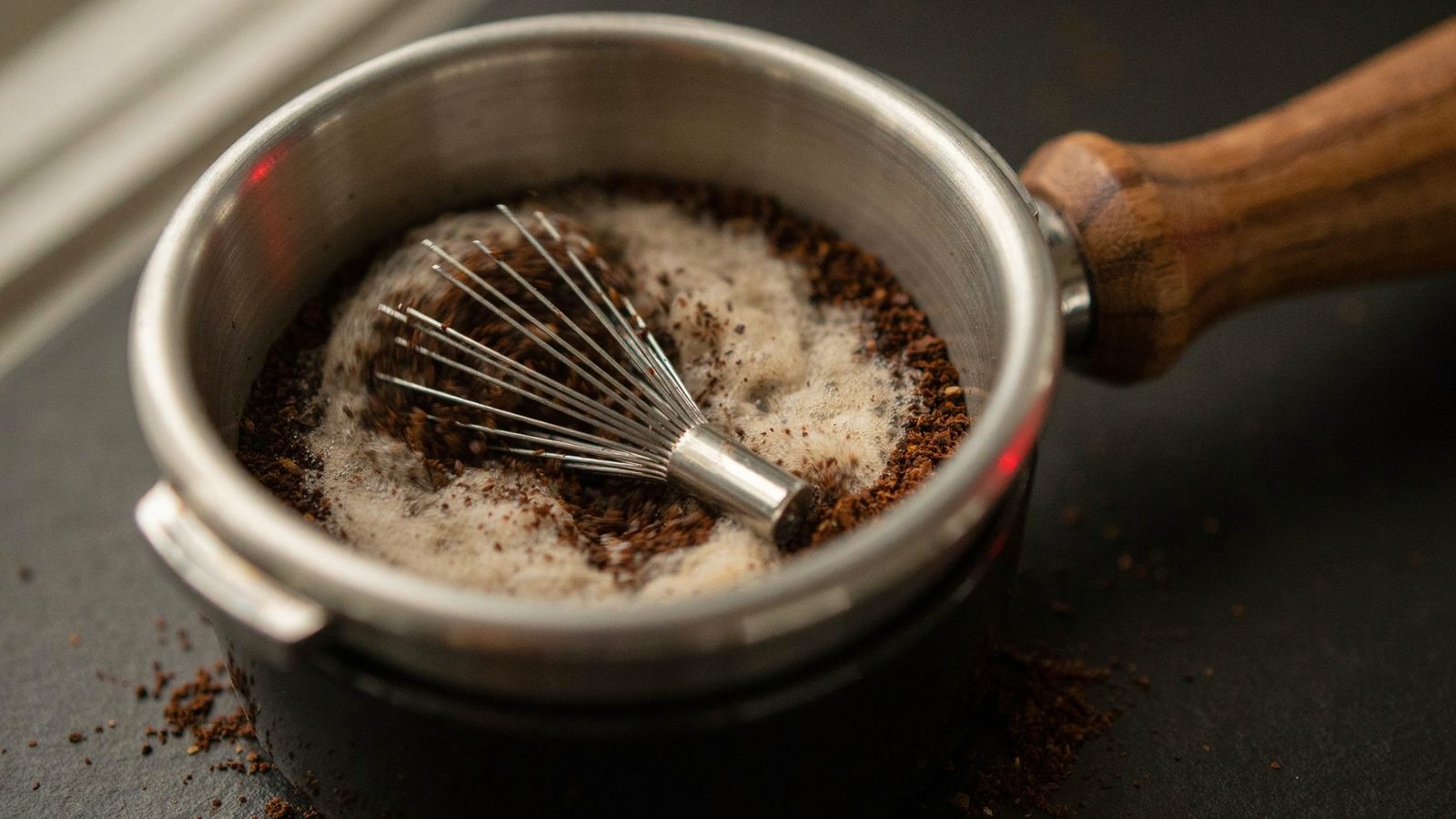 A WDT tool with fine stainless needles stirring fluffy freshly ground coffee inside an espresso basket, a walnut handle, crumbs of coffee on a dark mat