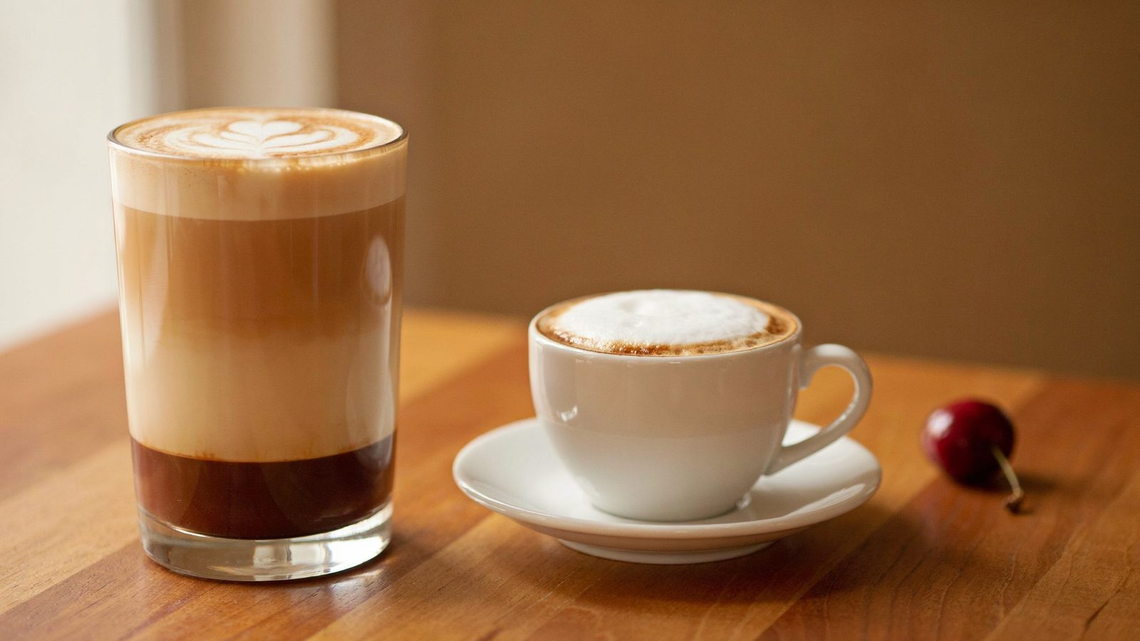 A latte in a tall glass and a cappuccino in a white ceramic cup side by side on a wooden table, the cappuccino with a thick foam cap and the latte with a thin layer and heart latte art